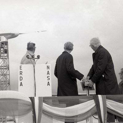 Fem menn står på et podium foran en vindturbin. To av mennene trykker på et teknisk apparat