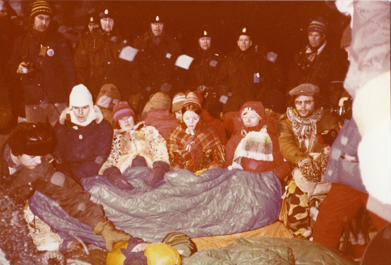 Demonstranter sitter med soveposer over seg, med politiet stilt opp bak.