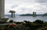 To oljeplattformer i betong under bygging i en fjord. i forgrunnen; taket på et hus, to røde biler og byggemateriell.  