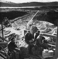 Fire menn i arbeidsklær spiser matpakke. De sitter på høyballer. I bakgrunnen er det en industritomt, bebyggelse, vann og fjell. 