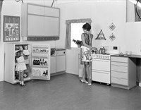 Kvinne i kjole og forkle foran komfyr. Liten jente i kjole står foran åpent kjøleskap. 1960-talls interiør i kjøkken. 